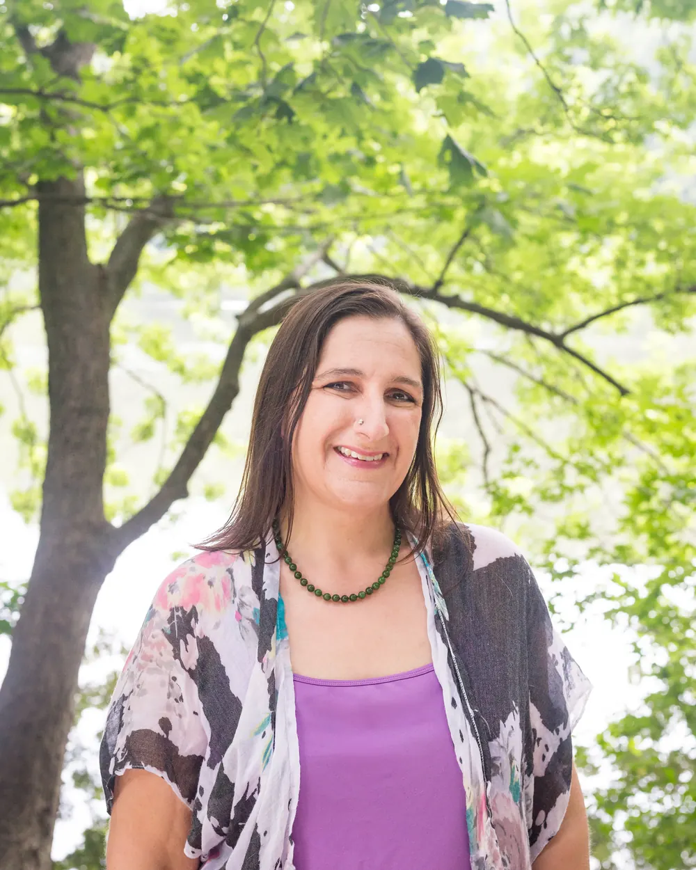 Kara Katz outdoors under green trees, smiling, in a lavender top and patterned cardigan