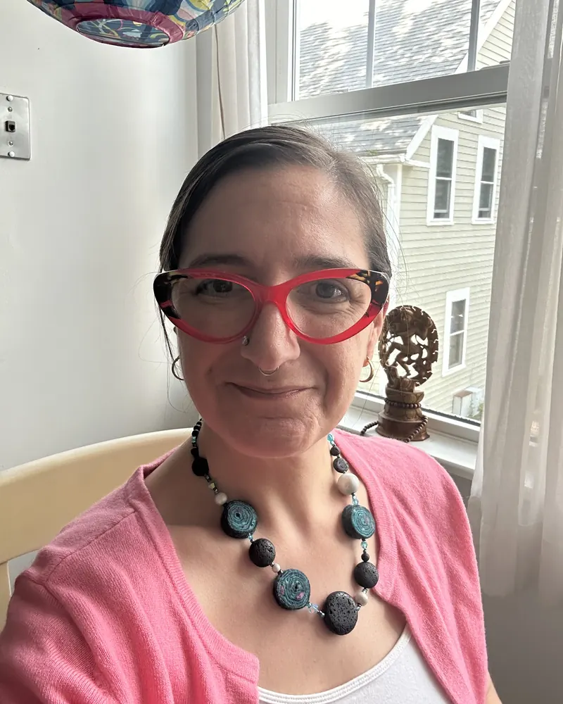 Kara Katz smiling by a sunny office window, wearing red glasses, a pink cardigan and a bold beaded necklace